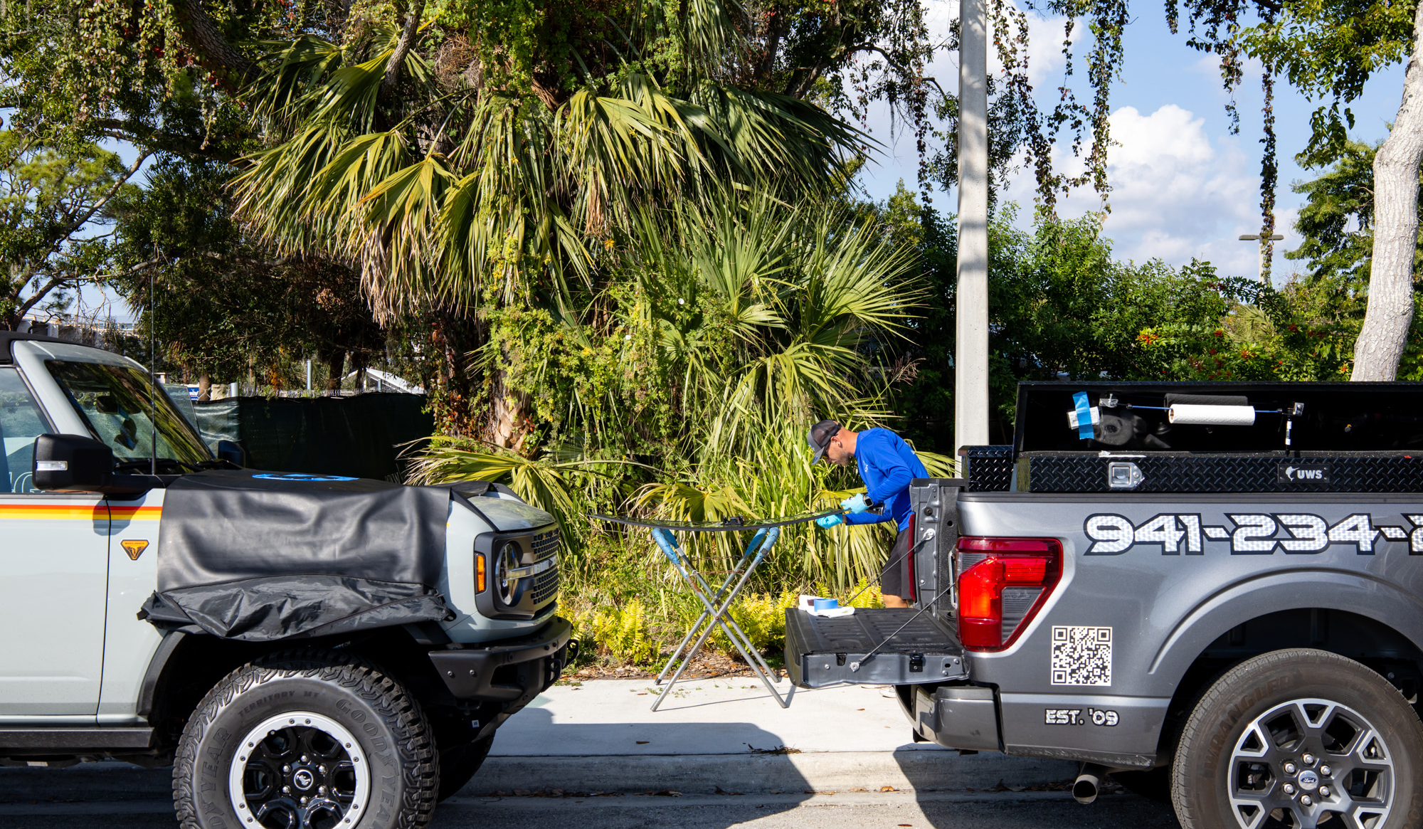 Auto glass technician replacing windshield in Florida, surrounded by palm trees and parked vehicles, showcasing mobile auto glass service.
